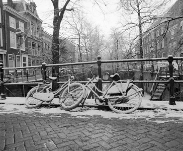 855683 Afbeelding van twee besneeuwde fietsen aan de reling op de Paulusbrug over de Nieuwegracht te Utrecht.